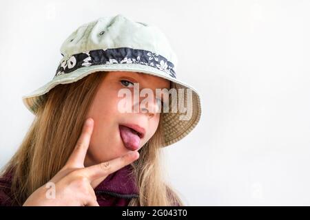 Das Mädchen streckte ihre Zunge aus und zeigt zwei Finger. Fröhliches Mädchen mit langen Haaren in einem grünen Hut. Vor der Kamera posieren. Die Freuden eines Kindes Stockfoto