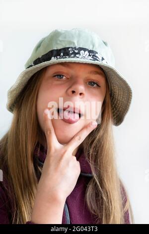 Das Mädchen streckte ihre Zunge aus und zeigt zwei Finger. Fröhliches Mädchen mit langen Haaren in einem grünen Hut. Vor der Kamera posieren. Die Freuden eines Kindes Stockfoto