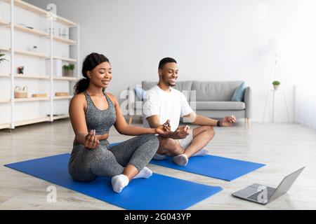 Glückliches schwarzes Paar praktiziert Yoga in der Nähe von Laptop, schaut sich Online-Video-Tutorial an, meditiert oder macht Atemübungen Stockfoto