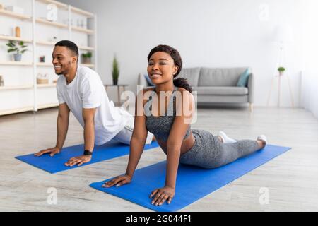 Cooles schwarzes Paar, das Kobra auf einer Yogamatte im Wohnzimmer posiert. Konzept für „Stay Home“-Aktivitäten Stockfoto