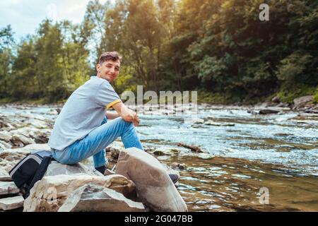 Junger Mann, der sich am Bergfluss entspannt und die natürliche Landschaft genießt. Reisender Rucksacktourist sitzt auf Felsen. Sommerurlaub Stockfoto