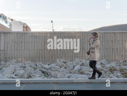 Hübsche und stilvolle Frau mittleren Alters, die ihr Gesicht abzieht Maske, die mit einer Mauer aus Ziegelsteinen durch die Stadt läuft Und Steine im Hintergrund Stockfoto