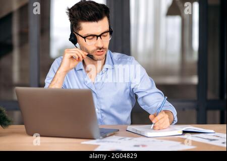 Fokussierter intelligenter erfolgreicher junger kaukasischer Geschäftsmann, Freiberufler oder Berater mit Brille und Kopfhörer, im Büro sitzend, mit Laptop, online mit dem Kunden chattierend, Notizen zu machen Stockfoto