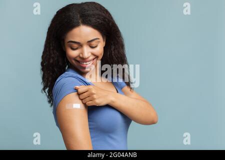 Werbung für die Impfung gegen Coronaviren. Glücklich geimpfte schwarze Dame zeigt Arm mit Gips auf blauem Hintergrund Stockfoto