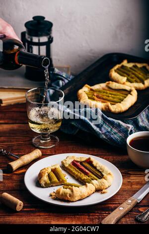 Mini-Galette mit Rhabarber-Scheiben auf weißem Teller. Hand mit Flasche Gießen Weißwein Stockfoto