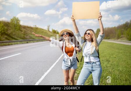 Zwei Freundinnen hitchen freie Fahrt, halten leeres Pappschild mit Mockup, halten Auto am Straßenrand, kopieren Raum Stockfoto