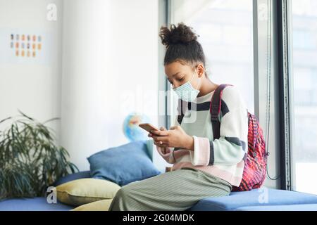 Seitenansicht Porträt eines afroamerikanischen Mädchens im Teenageralter, das im Klassenzimmer eine Maske trägt und auf das Smartphone schaut, mit kovidierten Sicherheitsmaßnahmen Stockfoto