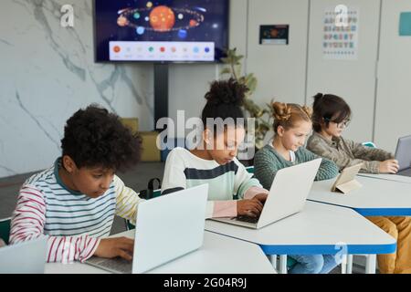 Eine vielfältige Gruppe von Kindern, die in der Schule in Reihe sitzen Und mit Computern Stockfoto