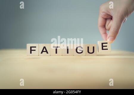 Die Hand legt einen Holzwürfel mit dem Buchstaben E aus dem Wort Müdigkeit. Das Wort steht auf Holzwürfeln, die auf dem Holztisch auf grauem Backgro stehen Stockfoto