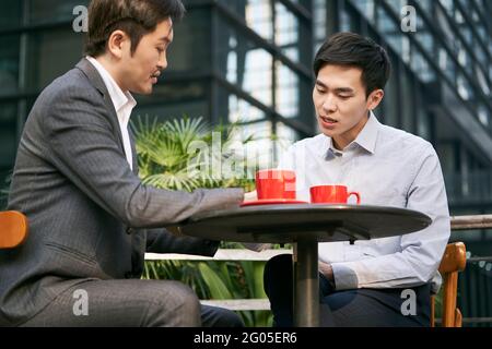 Zwei asiatische Geschäftsleute, die bei einem Kaffee im Freien über Geschäfte diskutieren Kaufen Stockfoto
