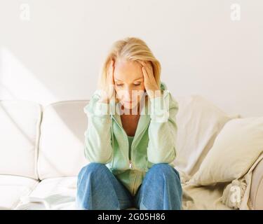 Müde oder melanikalisch reife 60s Frau in Casual Wear Gefühl aufgeregt, während sie auf dem Sofa mit Kopf in den Händen sitzt, traurige pensionierte Frau leidet unter Kopfschmerzen im Wohnzimmer. Depression nach der Pensionierung Stockfoto