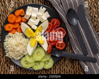 Dunkler Teller auf Strohmatte mit einfachem Reis, gehacktem gekochtem Ei, Kirschtomaten, geschnittenen Gurken, Karotten auf Rädern und griechischen Feta-Käsewürfeln Stockfoto