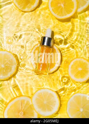 Luxus kosmetisches Öl oder Serum in Glasflasche mit Pipette in sauberem, transparentem Wasser mit Zitronenscheiben auf gelbem Hintergrund. Wasser spritzt im Sonnenlicht auf die Wasseroberfläche. Vertikaler Flatlay-Kopierbereich Stockfoto