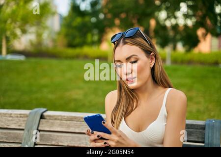 Die Frau in der Stadt sitzt im Sommer auf der Bank, hält Smartphone, liest und schreibt im Chat eine Nachricht, trägt Sonnenbrillen und Sommerkleidung. Hochwertige Fotos Stockfoto