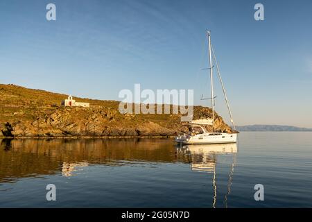 Alte, weiß getünchte verlassene Kirche Agios Ioannis auf einer dramatischen Klippe mit einem Segelboot, das in einer Bucht auf der griechischen Insel Kithnos vor Anker liegt, während es ruhig und warm danach war Stockfoto