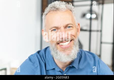 Kopfbild eines gutaussehenden, gut aussehenden, blauäugigen und reifen Mannes, der mit einem fröhlichen Lächeln auf die Kamera blickt. Nahaufnahme eines charismatischen grauhaarigen bärtigen älteren Mannes Stockfoto