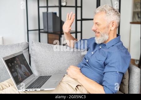 Zwei verschiedene männliche Kollegen sprechen über die Distanz via Videoverbindung. Älterer grauhaariger Mann begrüßt den afroamerikanischen Mann auf dem Laptop-Bildschirm. Reife Schüler lernen Sprache online Stockfoto