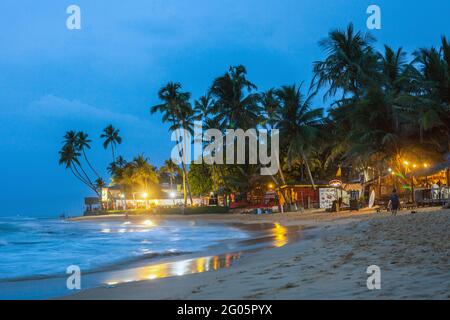 Strandszene mit Palmen und Hütten nach der Dunkelheit durch gelbe Straßenlaternen beleuchtet, Hikkaduwa, Südprovinz, Sri Lanka Stockfoto