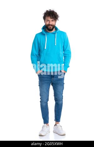 Lockiger, unrasierte Mann mit blauem Sweatshirt, der die Hände in die Jeanstaschen hält und lächelt, während er im Studio selbstbewusst vor weißem Hintergrund posiert Stockfoto