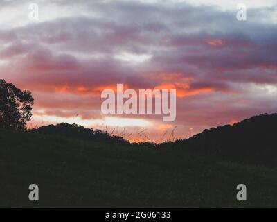 Pink Sunset, die untergehende Sonne, die die Wolken zu einem feurigen orangefarbenen Schein und grauer Purpurfarbe erhellt, Hügel darunter Stockfoto