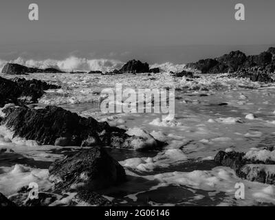 Einfarbiges, schaumiges Wasser in der Brandung, das nach rauem Wind durch die Felsen auf den Strand spült Stockfoto