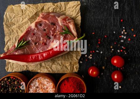 Rohes Stück Ribeye-Rindfleisch auf Bastelpapier auf einem Holztisch gestapelte Tomaten, garniert mit scharfem Paprika, Gewürzen und himalaya-Salz. Frisches Essen. Stockfoto