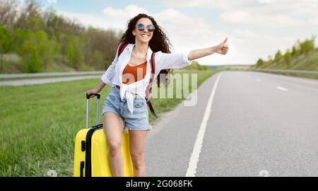 Die junge Dame, die ihre Fahrt mit dem Koffer auf der Autobahn abhing, versuchte, vorbeifahrende Autos zu fangen und auf dem Land zu trampeln Stockfoto