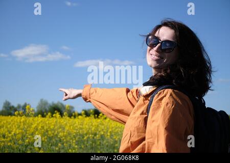 Das Mädchen in der Brille zeigt mit der Hand auf die Blumen. Reisen Sie außerhalb der Stadt. Sonniges Wetter. Wiese mit Blumen Stockfoto
