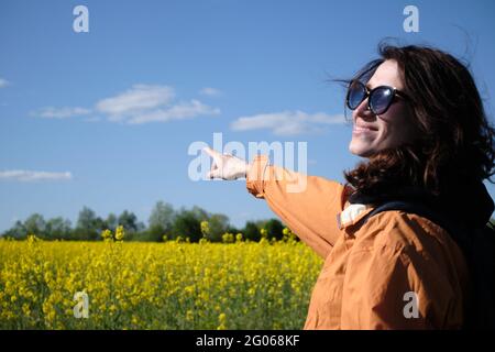 Das Mädchen in der Brille zeigt mit der Hand auf die Blumen. Reisen Sie außerhalb der Stadt. Sonniges Wetter. Wiese mit Blumen Stockfoto