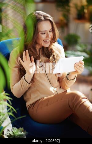 Green Home. Lächelnd trendy mittleren Alters Hausfrau mit langen welligen Haaren mit Tablet-PC in beige Hosen und Bluse mit Online-Meeting, während in einem sitzen Stockfoto