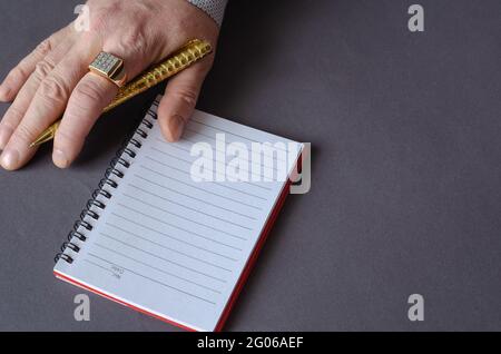 Eine Hand hält einen goldenen Stift vor einem grauen Hintergrund. Ein leeres offenes Notizbuch in der Nähe. Ein erwachsener Mann mit einem Siegel auf seinem Zeigefinger. Geschäftskonzept. Anzeigen Stockfoto