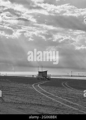 Ein dramatisch erleuchteter Blick auf einen fast leeren Margate-Strand, an dem Sonnenlicht durch die Wolke bricht, um eine Rettungsschwimmerhütte auf dem frisch gerackten Sand zu suchen. Stockfoto