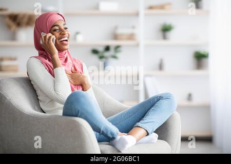 Fröhliche Schwarze Muslimische Frau In Hijab Im Gespräch Auf Dem Handy Zu Hause Stockfoto