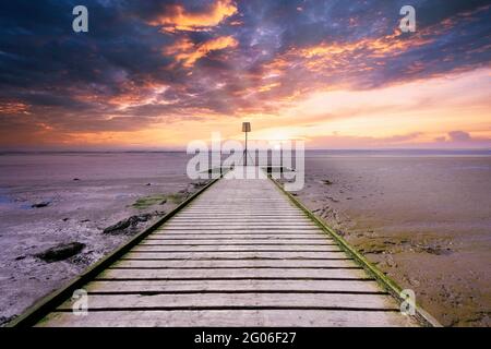 Die hölzernen Planken eines Steges in Lytham in Lancashire, Großbritannien, erstrecken sich über den Sandstrand, während die Sonne untergeht und einen dramatischen Himmel erzeugt Stockfoto