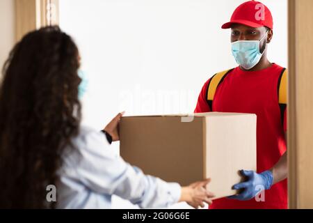 Schwarzer Liefermann in Maske, der der Frau einen Karton gab Stockfoto