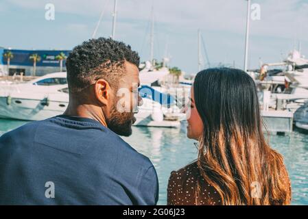 Junges multiethnisches Paar, das sich von hinten mit Booten im Hintergrund am Hafen ansieht Stockfoto