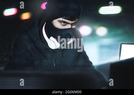 Wütender Computerhacker verschleierte Balaclava, die Daten über den PC aus seinem Underground Hideoutvor schwarzem Hintergrund und blauem Licht stiehlt. Porträt schließen. H Stockfoto