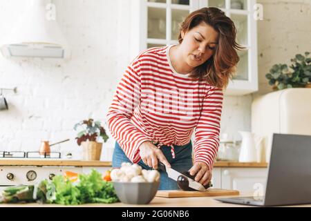 Attraktive junge Frau plus Größe Körper positiv in roten Langarm Kochen Lebensmittel mit Laptop mit Online-Video-Rezepte in der Küche zu Hause Stockfoto