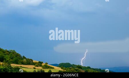 Gewitter über der hügeligen Landschaft. Blitzschlag trifft durch eine niedrige fliegende Wolke. Stockfoto