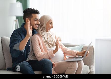 Fröhlich Schwanger Muslim Paar Mit Laptop Zu Hause Für Videoanruf Stockfoto