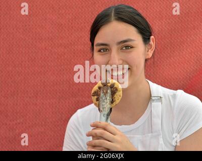Junge charmante Brünette Mädchen lächelt mit den Zähnen und hält mit Essenzangen ein hausgemachtes Gourmet-Schokoladenkekchen vor einem roten Hintergrund. Stockfoto