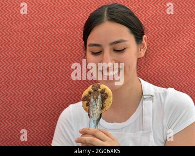 Junge charmante Brünette Verkäuferin lächelt und schaut auf einen frisch gebackenen Gourmet-Chocolate-Chip-Cookie in einer Essenzange vor einem roten Hintergrund. Stockfoto