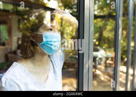 Traurige asiatische Frau, die Gesichtsmaske trägt und aus dem Fenster schaut Stockfoto