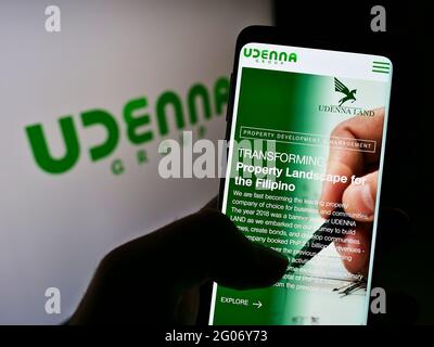 Person, die das Mobiltelefon mit der Webseite des philippinischen Konglomerats Udanna Corporation vor dem Logo auf dem Bildschirm hält. Konzentrieren Sie sich auf die Mitte des Telefondisplays. Stockfoto