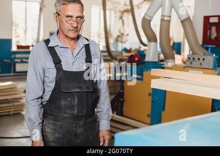 Porträt eines älteren Zimmermanns oder Zimmermanns in Overalls, der in einer Tischlerwerkstatt mit Holzbrettern arbeitet. Sägen von Holz. Stockfoto