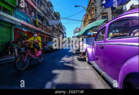 Eine Radfahrerin fährt an einem geparkten alten, violetten VW-Käfer vor dem Hintergrund farbenfroher Gebäude in der Chinatown-Gegend von Bangkok, Thailand, vorbei Stockfoto