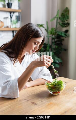 Enttäuschte junge Erwachsene Frau im Bademantel essen Gemüsesalat in der modernen Küche