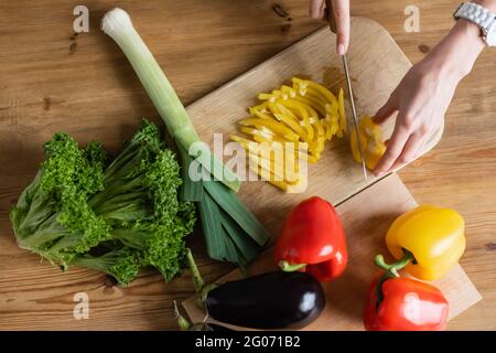 Draufsicht auf weibliche Hände, die in der Küche gelben Pfeffer auf dem Schneidebrett hacken und anderes Gemüse auf dem Tisch haben Stockfoto