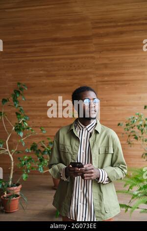 Afrikanischer junger Geschäftsmann in Brillen mit Mobiltelefon, während er im modernen Büro steht Stockfoto
