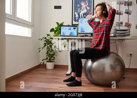 Frau mit Telearbeit, die vor ihr auf einem Fitball sitzt Schreibtisch, der über ein Headset spricht Stockfoto